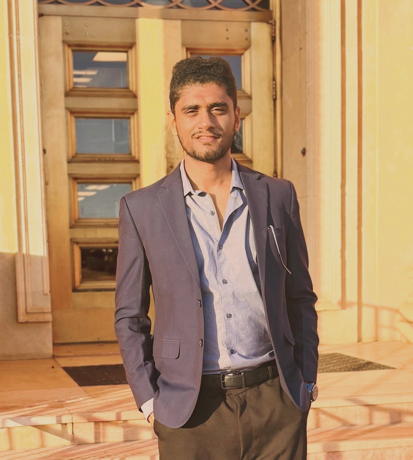 Arnav Chudiwale in a navy blazer, standing at the entrance of a stone building in evening light